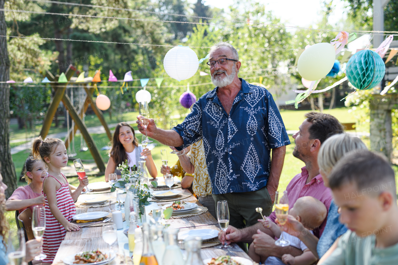 Grandfather clinking with champagne glass. Senior man standing, making celebratory toast at outdoor summer garden party.