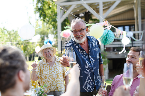 Grandfather clinking with champagne glass. Senior man standing, making celebratory toast at outdoor summer garden party.