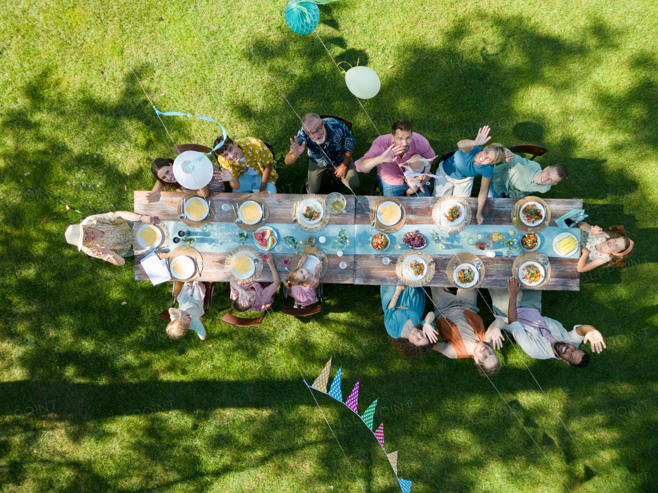 Top view of people at summer garden party in a beautiful garden. BBQ family gathering, with a set table and colorful decorations. Family waving up.