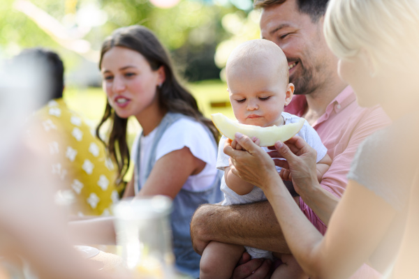 Cute baby eating honeydew at a summer garden party. The father is holding the baby boy in his arms, mother feeding him.