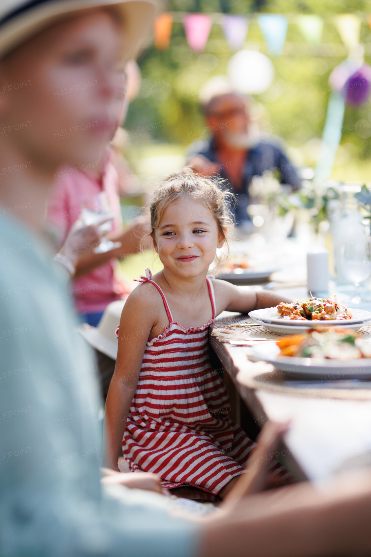 Portrait of a beautiful little girl sitting at a table eating grilled food outdoors. Girl at family garden party.