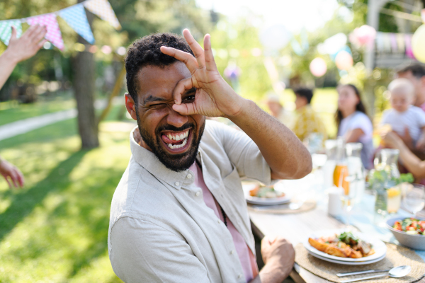 Portrait of handsome man making funny faces, sitting at the party table during a summer garden party with family and friends.