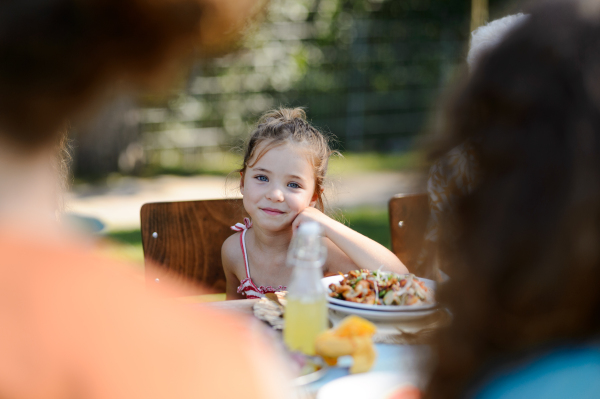 Portrait of a beautiful little girl sitting at a table eating grilled food outdoors. The girl at family garden party.