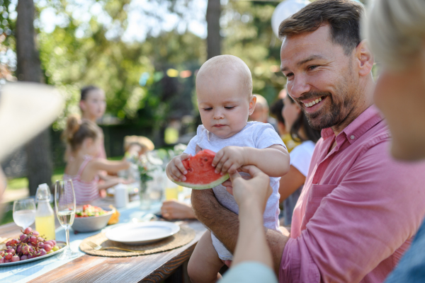 Cute baby eating watermelon at a summer garden party. Handsome father holding the baby boy in his arms and smiling.