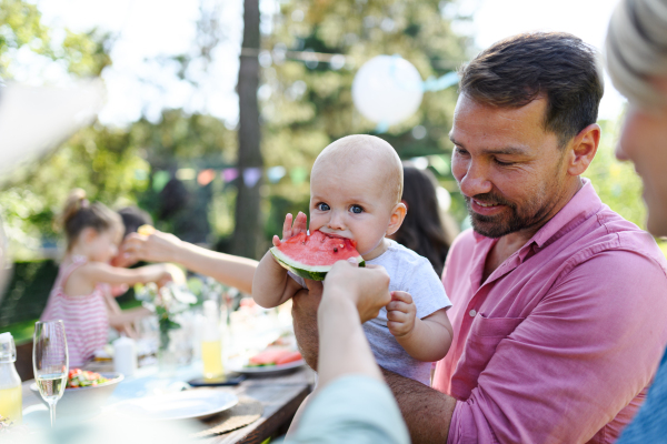 Cute baby eating watermelon at a summer garden party. The father is holding the baby boy in his arms and smiling.