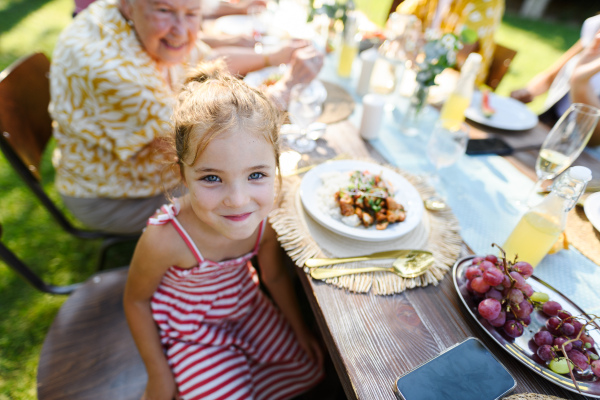 Portrait of a beautiful little girl sitting at a table eating grilled food outdoors. Girl at family garden party.