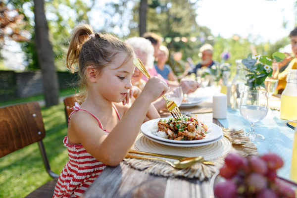 Portrait of a beautiful little girl sitting at a table eating grilled food outdoors. Girl at family garden party.