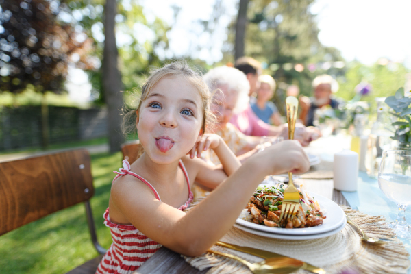 Portrait of a funny little girl sitting at table eating grilled food outdoors, sticking out her tongue. Girl at family garden party.