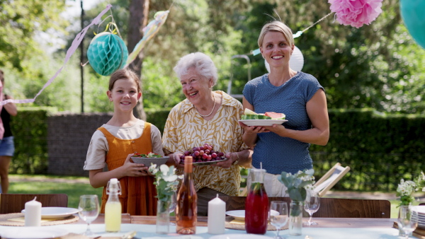 Grandmother, mother and daughter setting table for summer garden party. Three generations of women in family, bringing plates, food, and drinks at table.