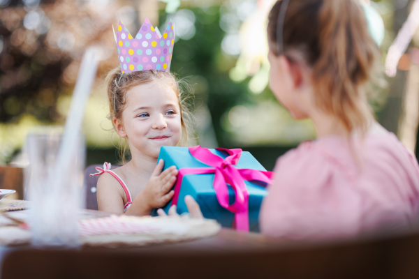 Little friends at a birthday party outdoors at garden. Cute girl with paper crown giving the birthday girl gift. Birthday garden party for children.