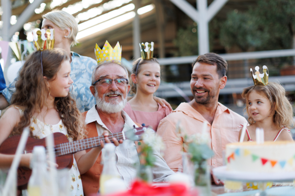 Family birthday garden party. Family sitting at the table having fun, little girl playing ukulele for the birthday man.