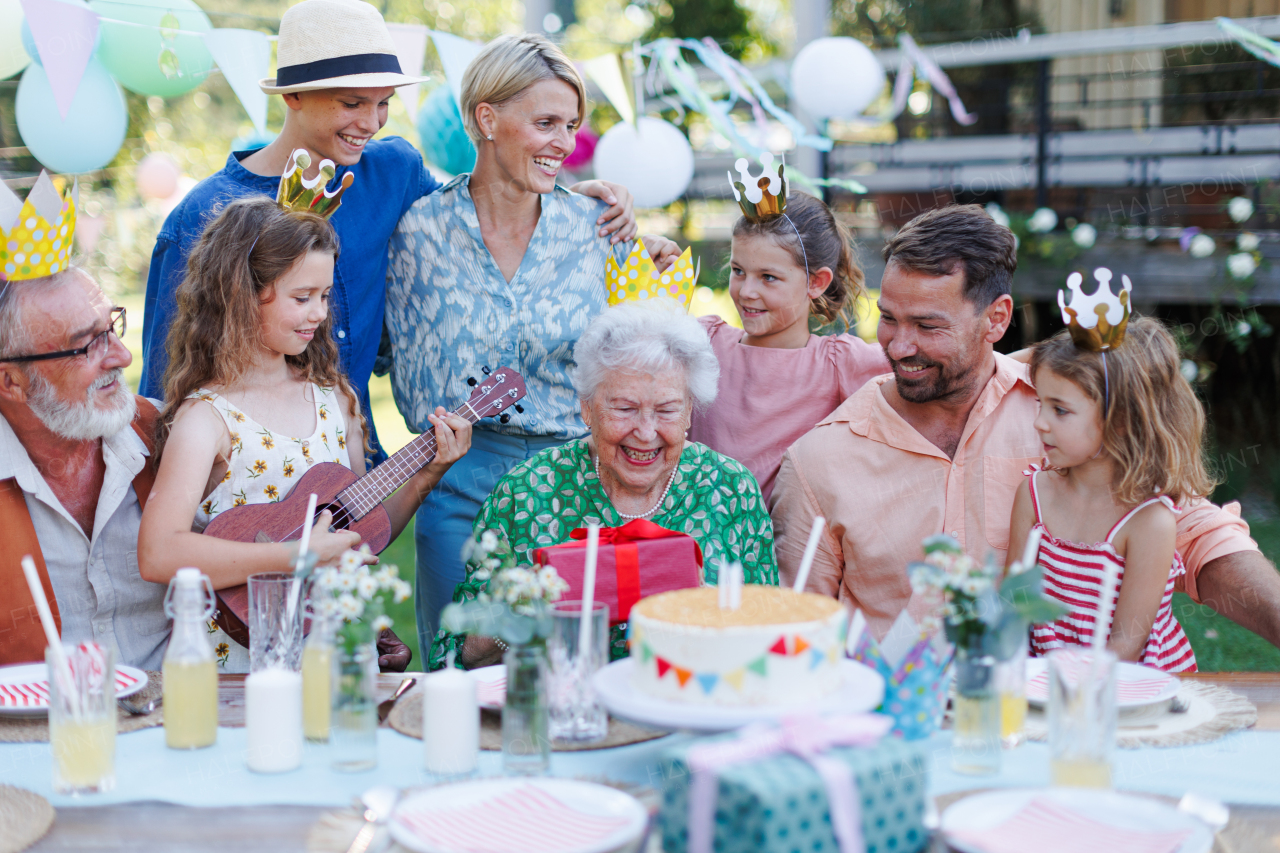 Garden birthday party for senior lady. Beautiful senior birthday woman receiving gift from granddaughter, family and friends. Birthday celebration with paper crowns.