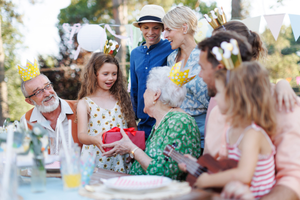 Garden birthday party for senior lady. Beautiful senior birthday woman receiving gift from granddaughter, family and friends. Birthday celebration with paper crowns.