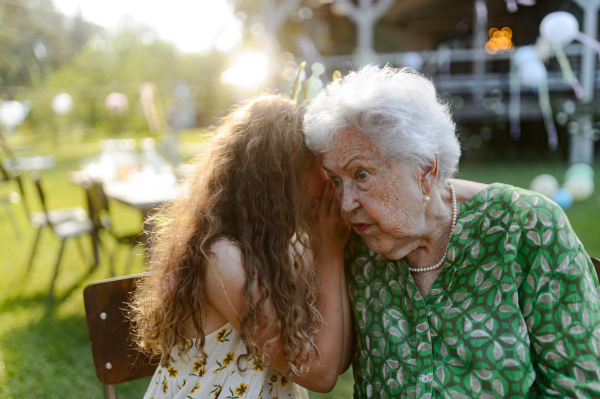 Young girl whispering a secret into grandmother's ear at a garden party. Love and closeness between grandparent and grandchild.