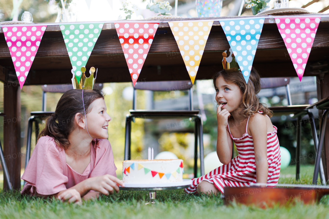 Cute girls with paper crowns eating birthday cake under table. Birthday garden party for children. Birthday garden party for children.