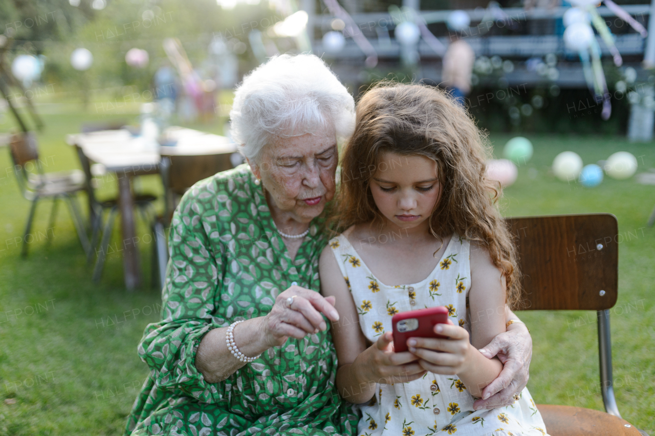 Young girl showing something on smartphone to elderly grandmother at a garden party. Love and closeness between grandparent and grandchild.