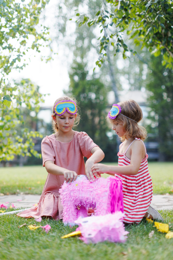 Two girls smashing and opening pink pinata with a stick at birthday party. Children celebrating birthday at garden party. Children having fun and playing.