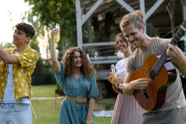 Handsome man playing at guitar for dancing friends at party. Friends and family dancing and having fun at a summer grill garden party.