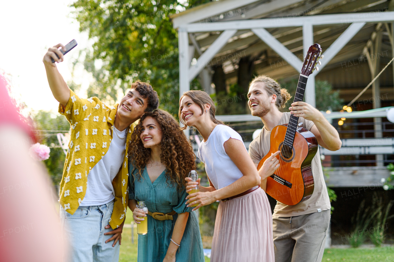 Friends taking selfie at a summer grill garden party. Handsome man playing at guitar at party.