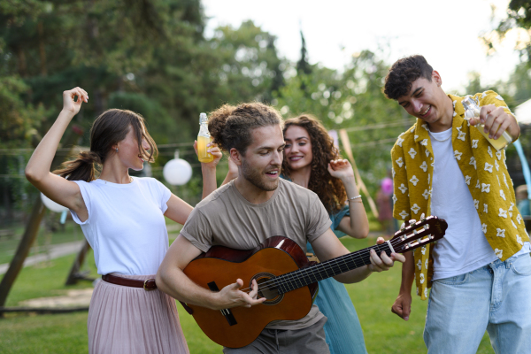 Handsome man playing at guitar for dancing friends at party. Friends and family dancing and having fun at a summer grill garden party.