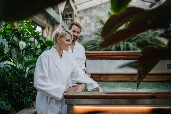 Beautiful couple standing by hot tub, wearing bathrobes, enjoying romantic wellness weekend in the spa. Concept of Valentine's Day.