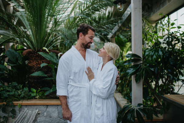 Beautiful couple in spa robes standing in hotel greenhouse, looking at each another, enjoying romantic wellness weekend in the spa. Concept of Valentine's Day.