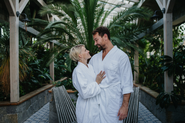 Beautiful couple in spa robes standing in hotel greenhouse, looking at each another, enjoying romantic wellness weekend in the spa. Concept of Valentine's Day.