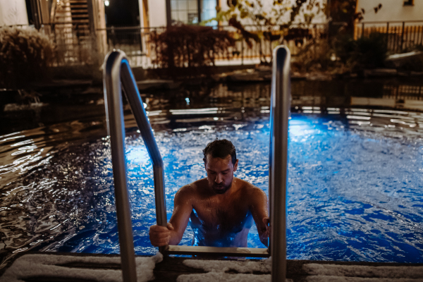 Mature man getting out of outdoor pool at night, enjoying calmness and empty pool. Wellness weekend in the hotel.