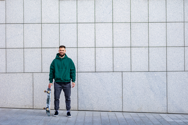 Stylish skateboarder standing in front building, looking at camera, smiling. Young man skating outdoors in the city. Concept of skateboarding as sport and lifestyle.