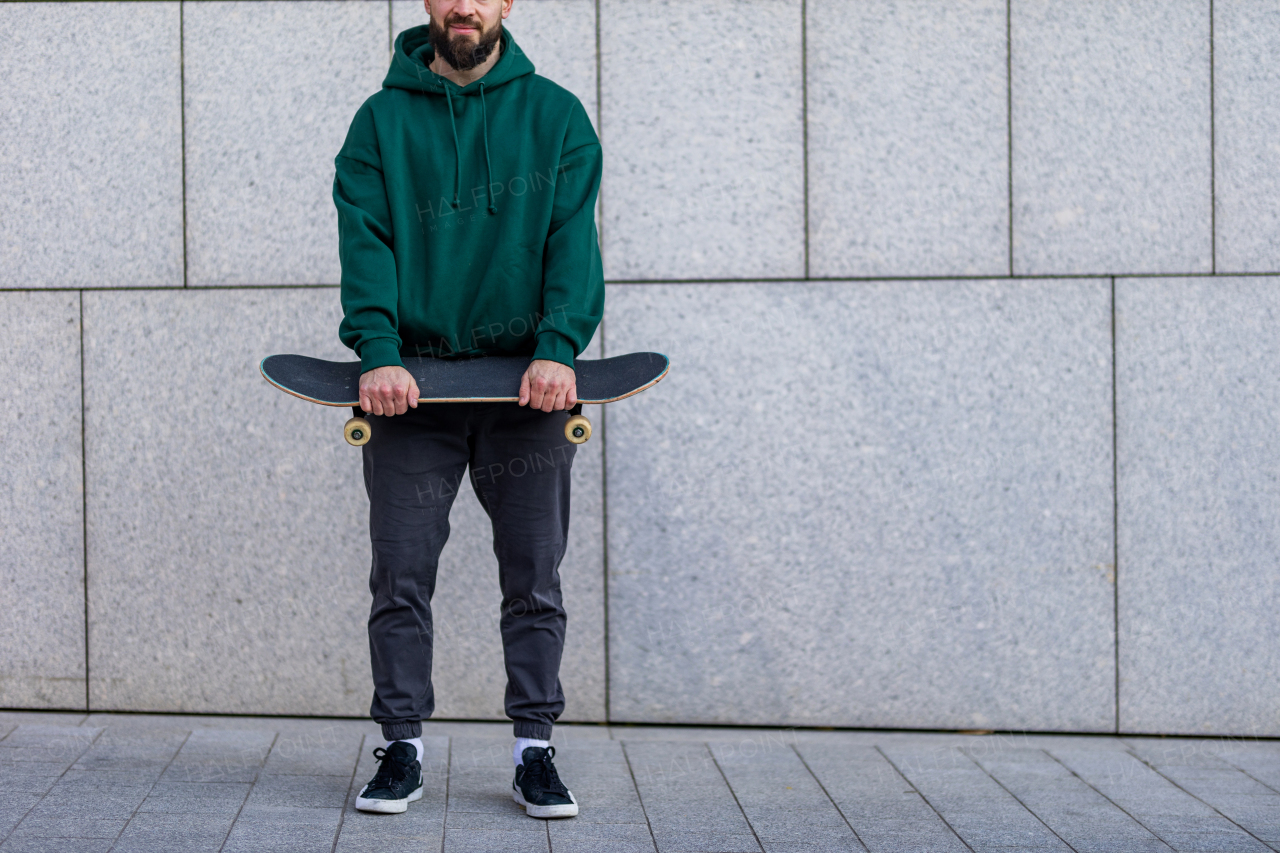 Stylish skateboarder standing in front building, looking at camera, smiling. Young man skating outdoors in the city. Concept of skateboarding as sport and lifestyle.