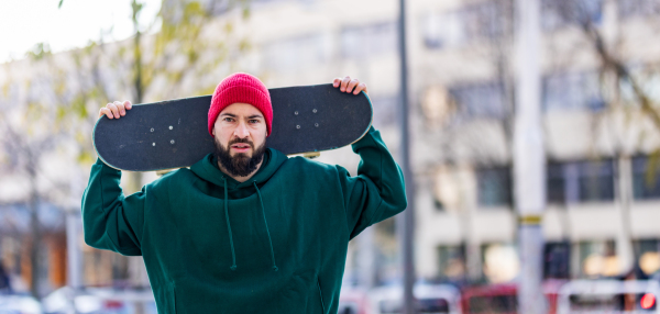 Portrait of stylish skateboarder with skateboard behind his head, looking at camera. Young man skating outdoors in the city. Concept of skateboarding as sport and lifestyle. Banner with copy space.