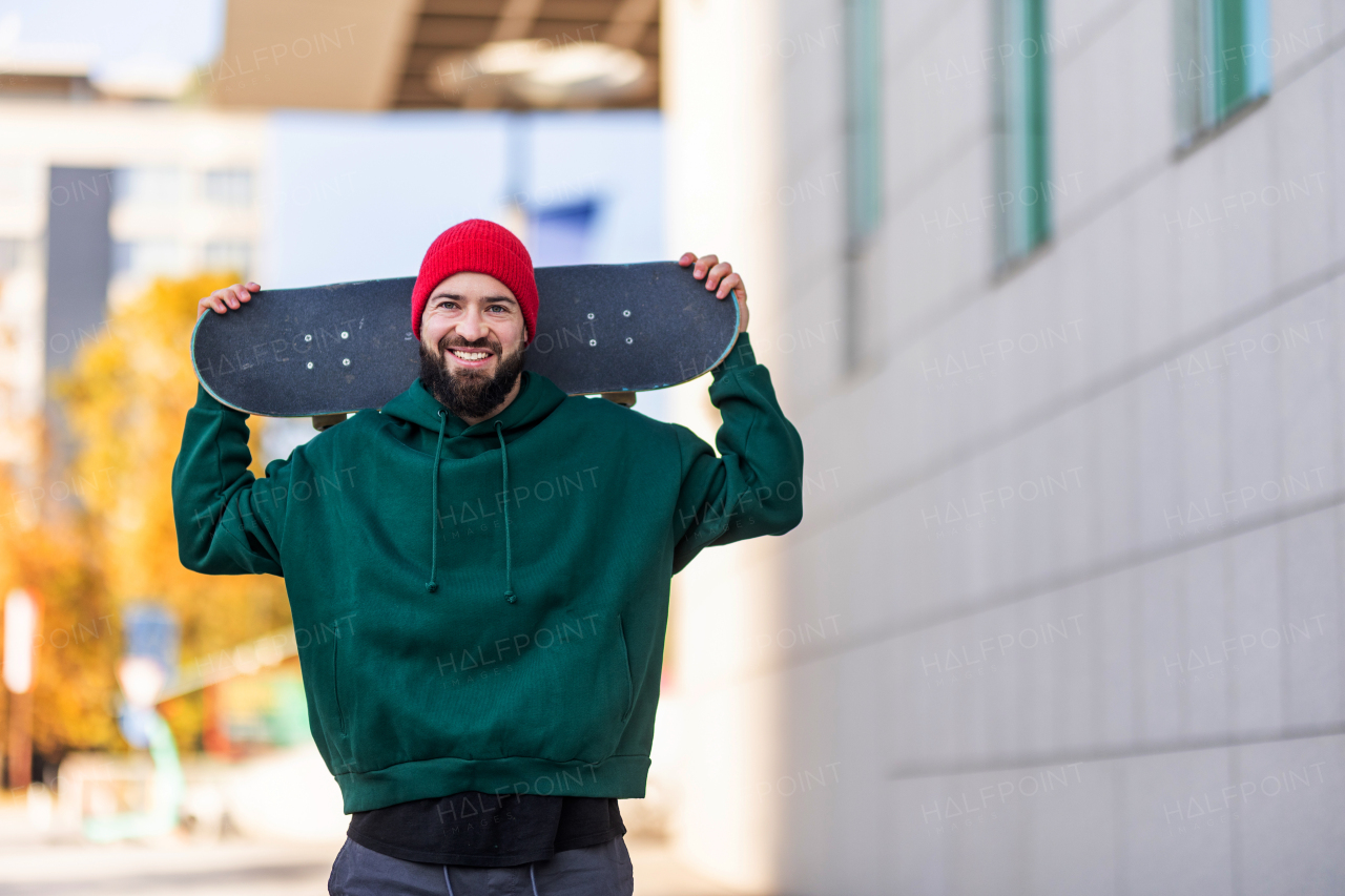 Portrait of stylish skateboarder with skateboard behind his head, looking at camera. Young man skating outdoors in the city. Concept of skateboarding as sport and lifestyle.