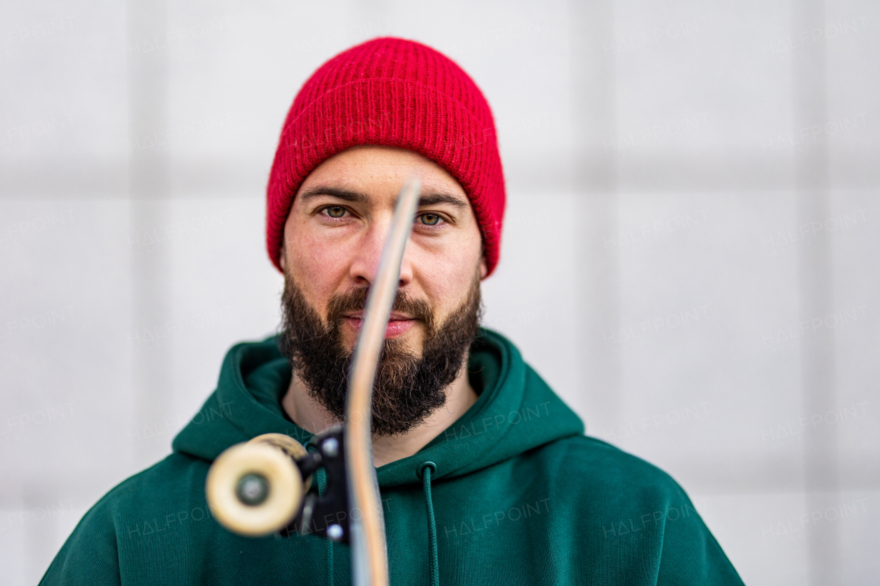 Portrait of stylish skateboarder with skateboard in front of face, looking at camera. Young man skating outdoors in the city. Concept of skateboarding as sport and lifestyle.