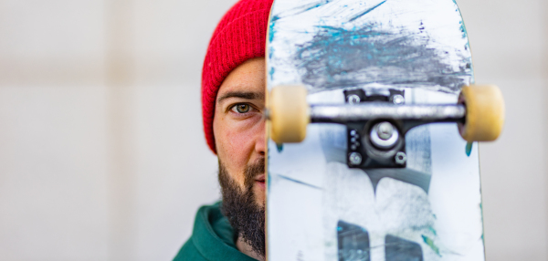 Portrait of stylish skateboarder with skateboard in front of face, looking at camera. Young man skating outdoors in the city. Concept of skateboarding as sport and lifestyle. Banner with copy space.