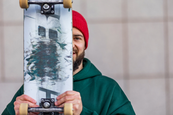 Portrait of stylish skateboarder with skateboard in front of face, looking at camera. Young man skating outdoors in the city. Concept of skateboarding as sport and lifestyle.