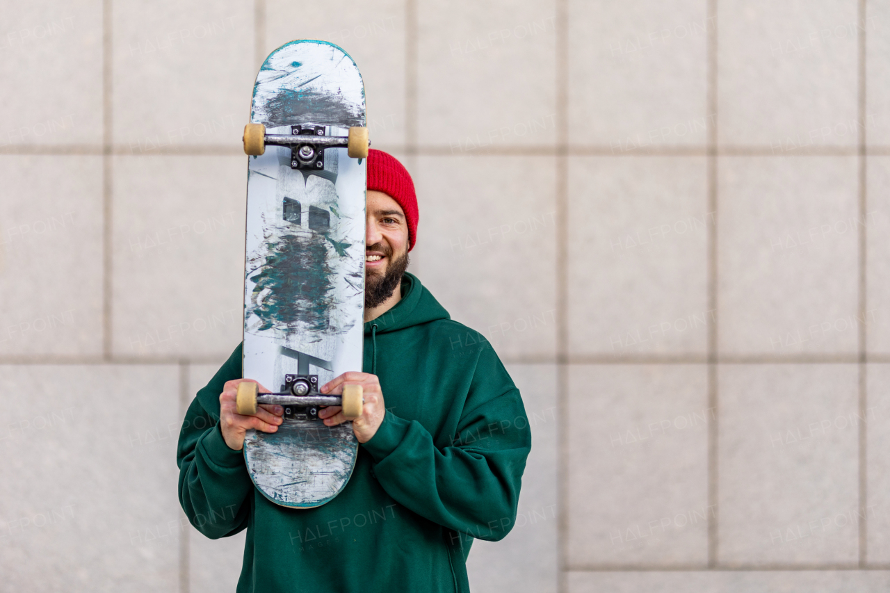 Portrait of stylish skateboarder with skateboard in front of face, looking at camera. Young man skating outdoors in the city. Concept of skateboarding as sport and lifestyle.