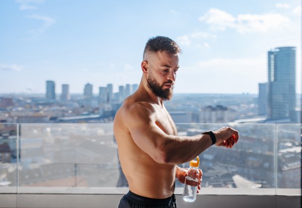 Man after home workout, checking his exercise performance on smartwatch, standing on balcony. Morning or evening workout routine.