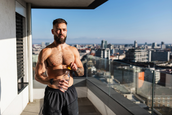 Handsome man with bare chest standing on balcony and enjoying beautiful view on the city. Relaxing after workout. Concept of selfcare and enjoying life for young people.