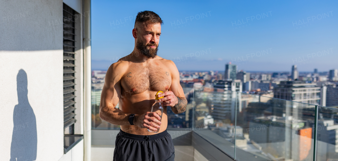 Handsome man with bare chest standing on balcony and enjoying beautiful view on the city. Relaxing after workout. Concept of selfcare and enjoying life for young people. Banner with copy space.