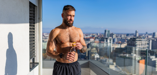 Handsome man with bare chest standing on balcony and enjoying beautiful view on the city. Relaxing after workout. Concept of selfcare and enjoying life for young people. Banner with copy space.