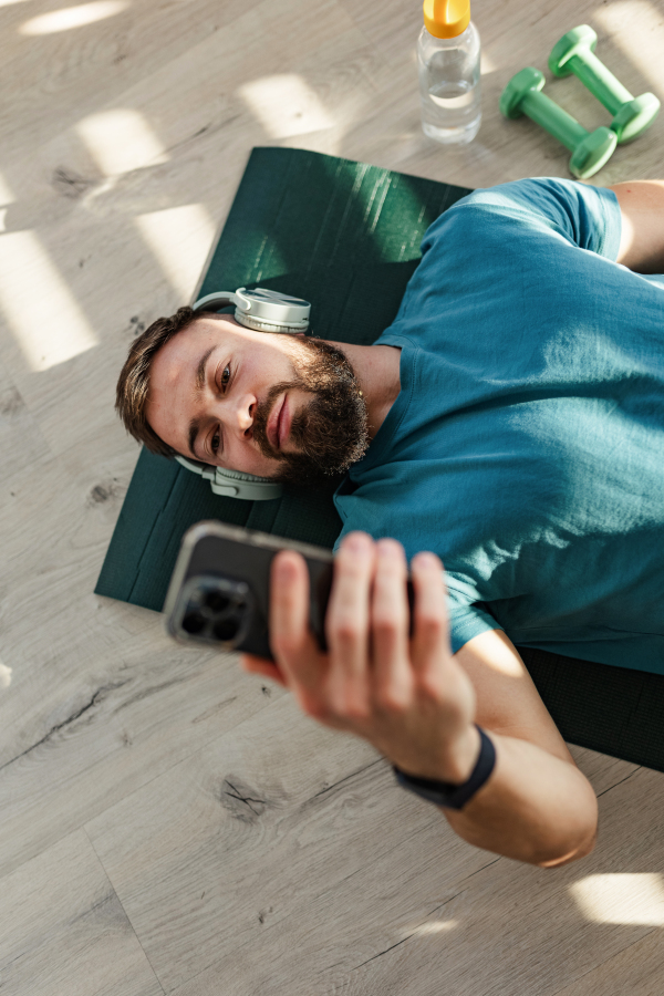 Young man resting after home workout, checking his exercise performance on smartwatch. Concept of morning or evening workout routine, listening to music through wireless headphones