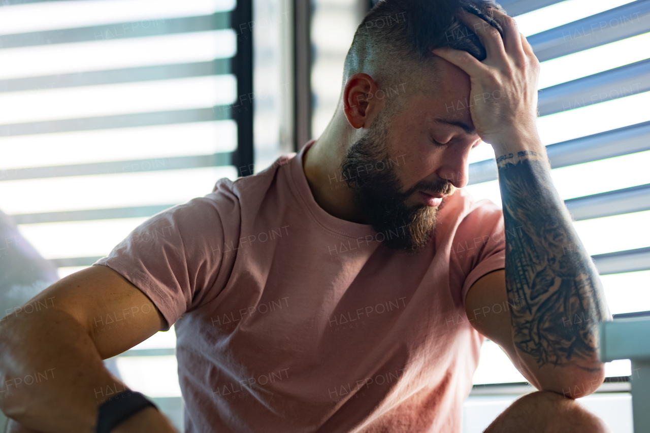 Anxious, depressed man in pink t-shirt with tattoo on arms, closed eyes, holding head in hand.