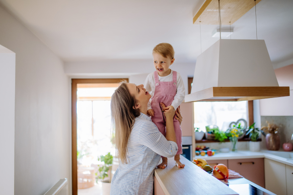 A young mother holding her little daughter when walking on kitchen counter at home.