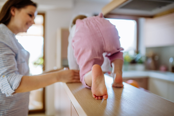 A young mother holding her little daughter when walking on kitchen counter at home. Rear view.