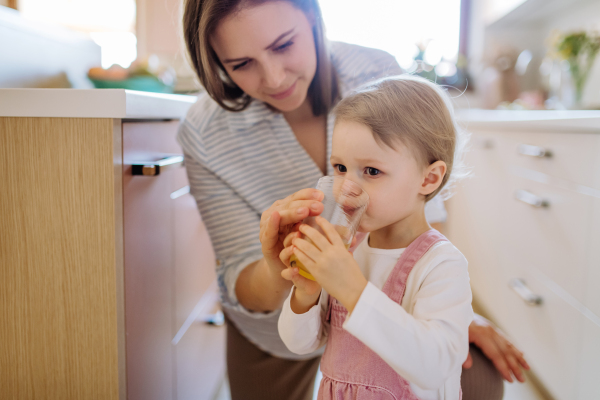 A young mother giving glass of water to drink to her little daughter in kitchen at home.