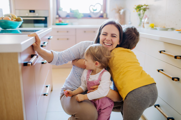 A young cheerful mother playing with her little children and having fun when hugging them in kitchen.