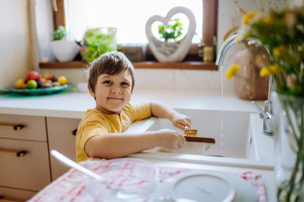 A little boy washing the dishes in sink in kitchen with wooden scrub, sustainable lifestlye. Looking at camera and smiling.