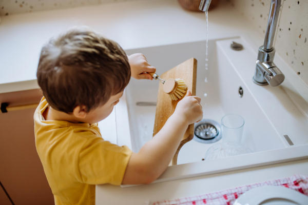 A little boy washing cup in sink in kitchen with wooden scrub, sustainable lifestlye.