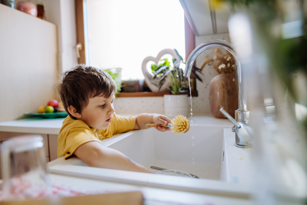 A little boy washing cup in sink in kitchen with wooden scrub, sustainable lifestlye.