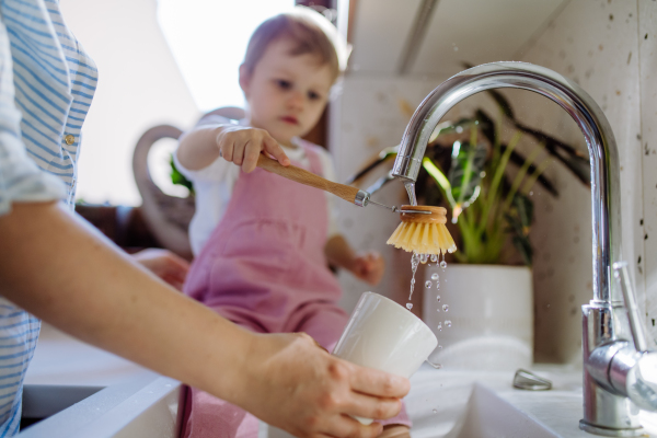 Little girl sitting on kitchen counter and helping mother to wash cup in sink in kitchen.